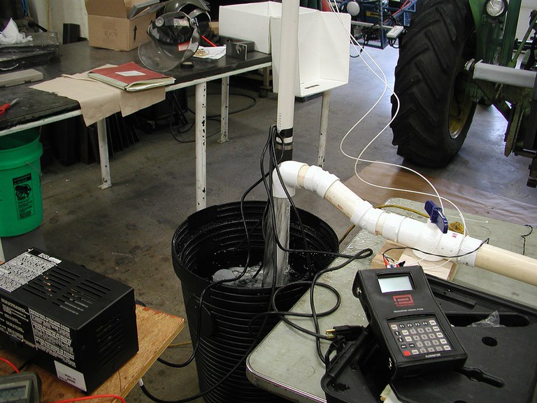 Figure 8. Backup pump test stand showing water flowing into sump that contains the two pumps. The charger is in the lower left and on the right is the ultrasonic flow meter used to measure the flow rate.