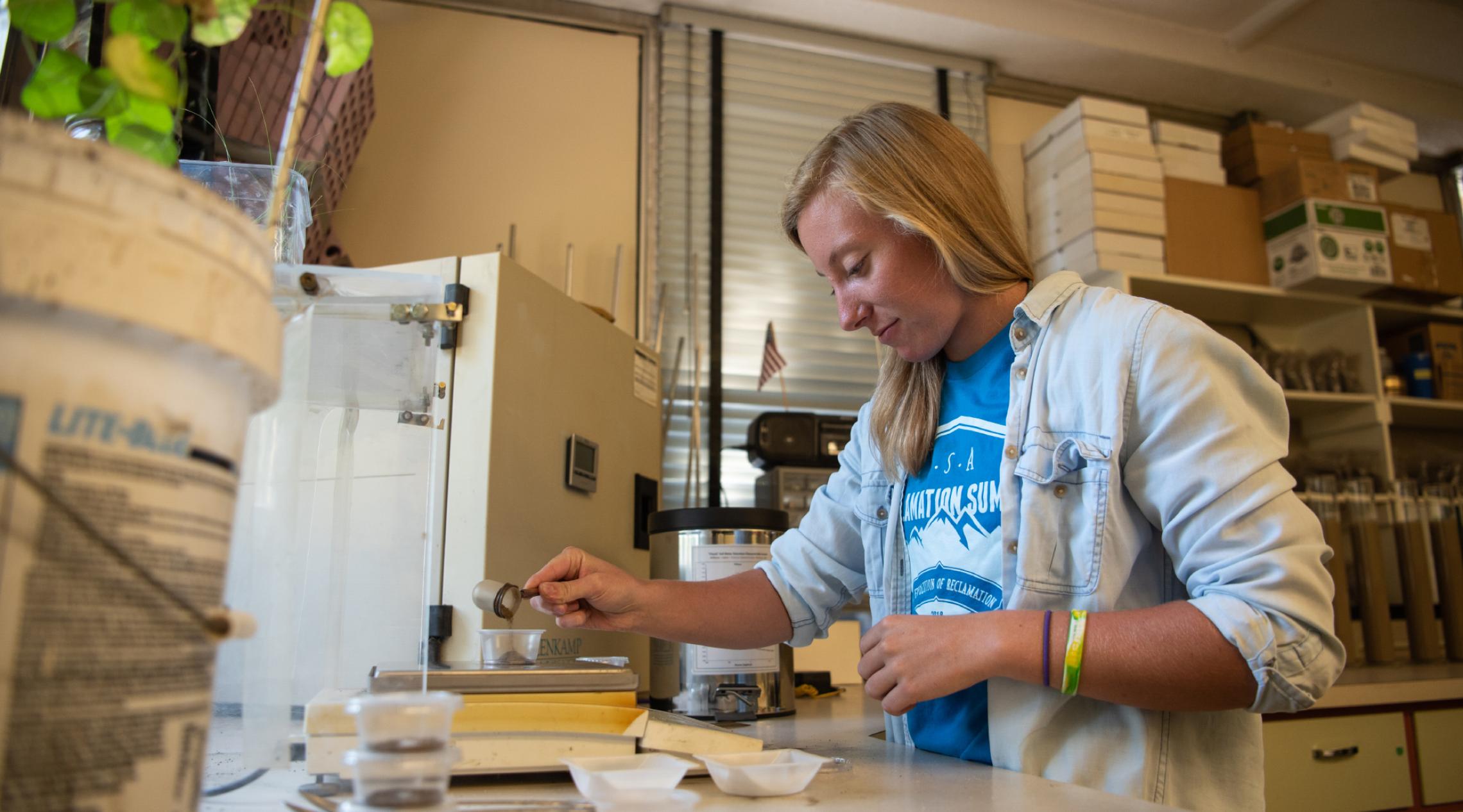 A young woman in a lab pours a small amount of soil into a clear plastic cup.