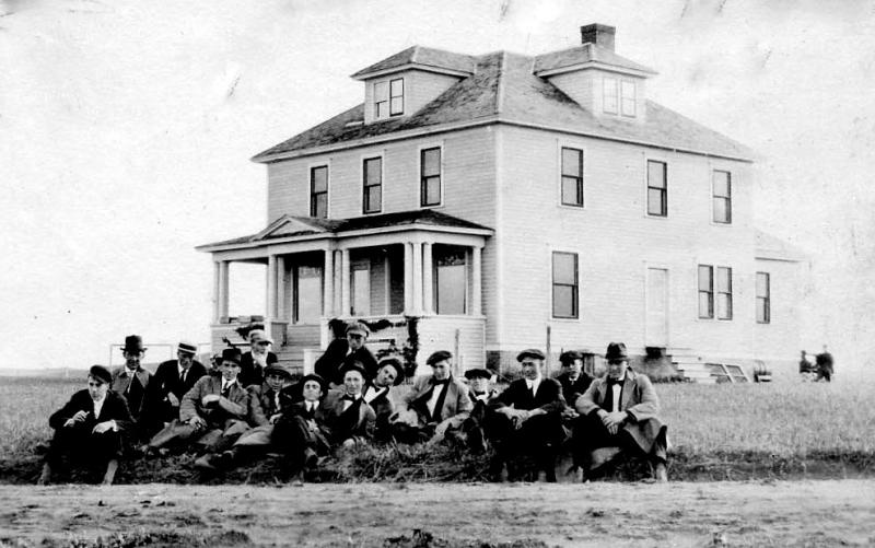 Black & white photo of the Hettinger REC house with dressed up young men sitting in front , 1913