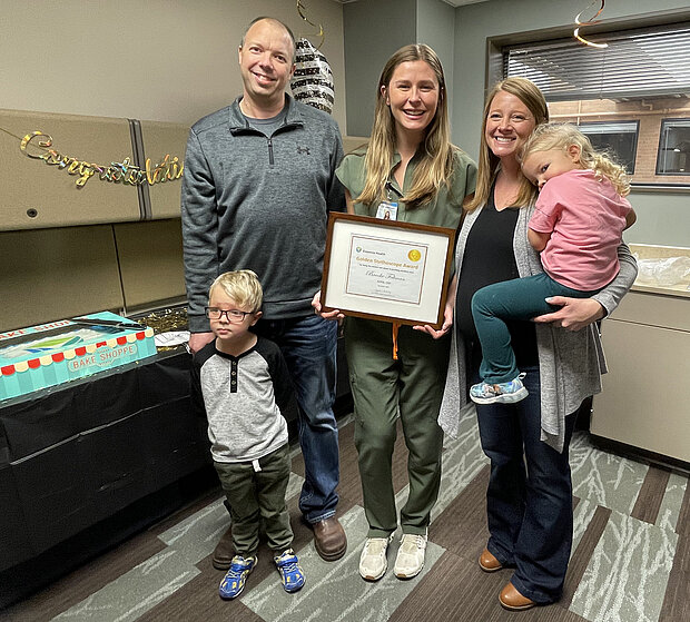 Photo of NDSU Nursing alum Brooke Feltman, DNP, holding a framed certificate, surrounded by patient's family that she helped, including a mother, father and two small children.