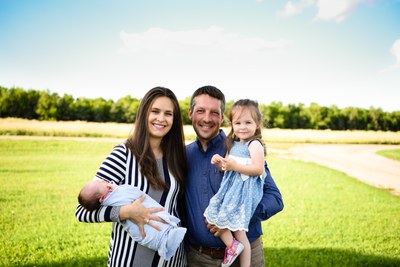 A family of four stand in in a farm yard. One parent is holding a toddler. The other is holding an infant. Green grass, a line of dark green trees and a blue sky with puffy white clouds are visible behind them.