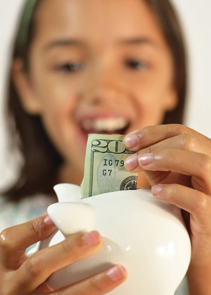 young smiling girl depositing a $20 bill into a small piggy bank 