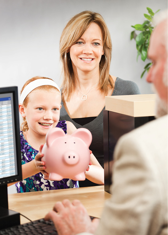 young girl in braces turning in a piggy bank to bank teller, with proud parent in background