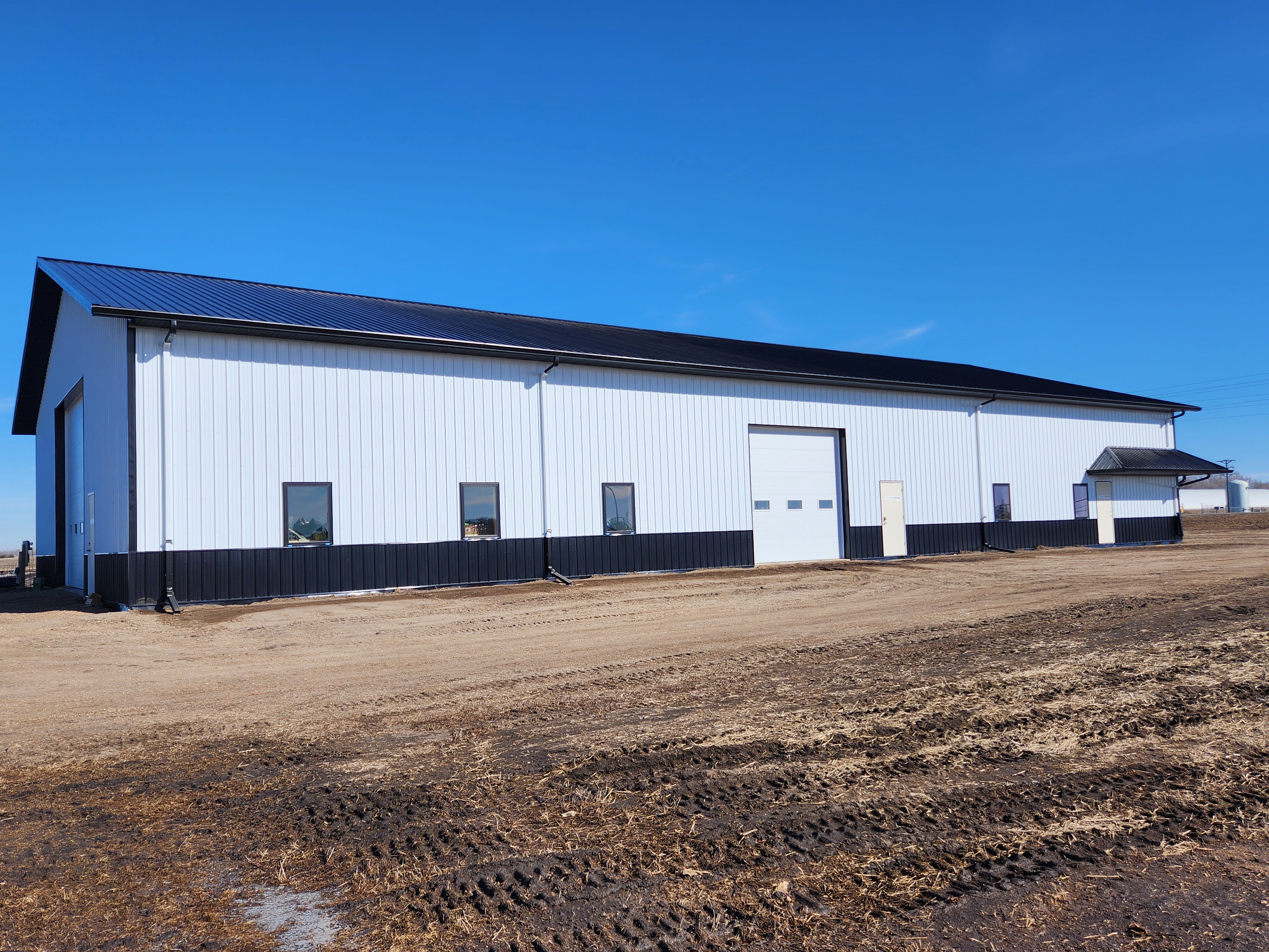 A 65-foot by 120-foot steel building with black wainscotting, white side walls, and a black roof stands on a gravel yard at the NDSU Oakes Irrigation Research Site. 