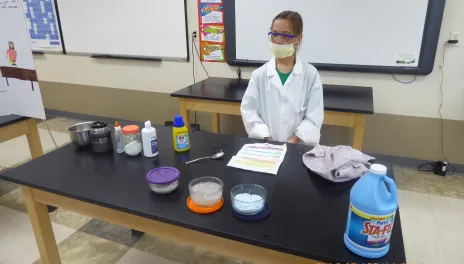 girl with mask and science lab coat on standing behind table with experiment ingredients