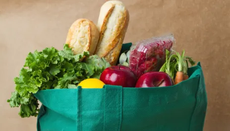 Food overflowing from reusable grocery tote, baguettes, fresh greens, apples, and carrots