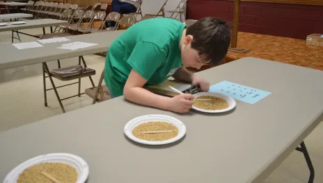 boy writing on sheet of paper with paper plates of seeds in front of him on table