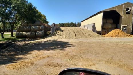 A large pile of corn silage beside a yellow, metal building. A stack of round hay bales and a line of large trees are visible on the other side of the pile. 