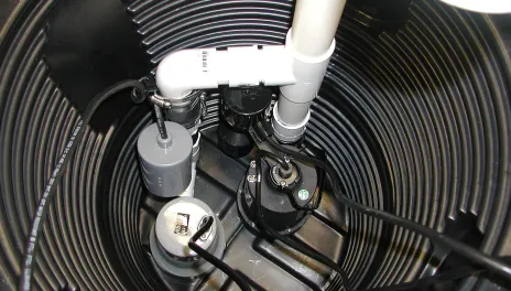 A black sump pump sits in a hole lined with black corrugated plastic. Electrical wires lead to the pump, connected to white PVC pipes rising from the hole.