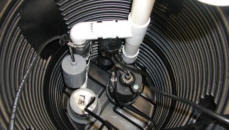 A black sump pump sits at the bottom of a hole lined with black corrugated plastic. Black electrical wires lead to the pump which is connected to white PVC pipes leading up out of the hole. 