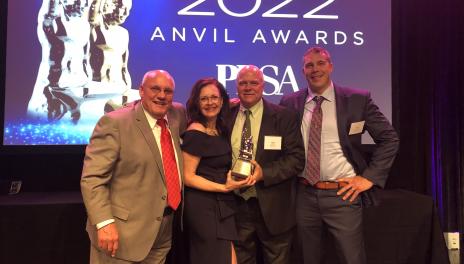 Four people stand on a stage in a line. the two people in the center are holding an award together. Behind them a screen displays the words "2022 Anvil Awards. PRSA."