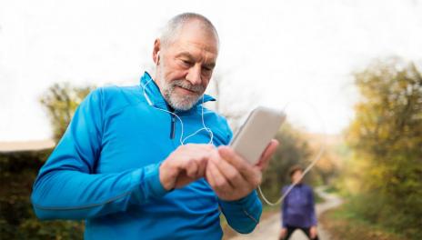 An older man standing on a running path looking at his mobile device. He's wearing athletic clothing and earbuds.