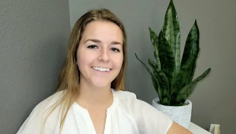 Female in white shirt smiling while standing next to a green plant. 