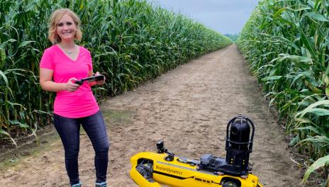 Woman in pink shirt stands with robot in corn field.