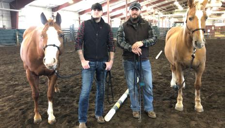 Two men pose with two horses in an arena