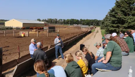 Two speakers lean on a feed bunk, talking to people on a flatbed trailer, with a dirt feedlot, red cattle, and a yellow polebarn in the background.
