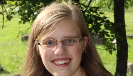 A smiling woman with long blonde hair, blue eyes and glasses, stands in front of a tree and green grass.