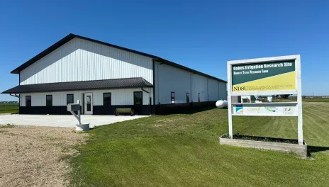 A large white building with green roof and trim sits at the Oakes Irrigation Research Site. A sign in the foreground labels the site.