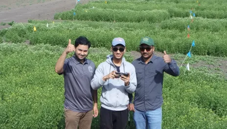 Three men stand together on bare ground ahead of several agronomy trial plots. The men on the left and right are giving a thumbs-up sign and the middle man is holding a small drone controller. 