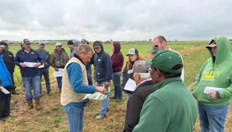 One man points to information on sheet of paper while he addresses a group of people in a field on a cloudy day.