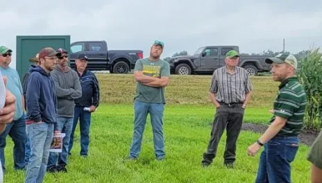 A group of people standing on a green field surrounding a presenter in front of tall corn plants and other forage crops.