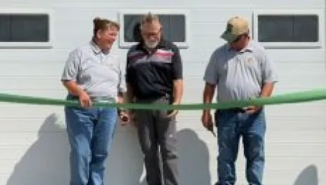 Three people, standing in front of a white garage door, are holding a green ribbon.