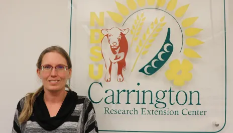 A woman in a black striped sweater stands in front of a graphic sign for the Carrington Research Extension Center.