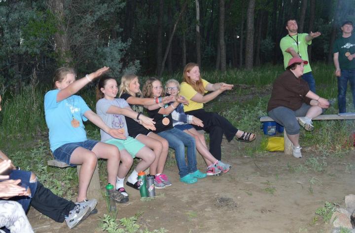 Youth and counselors in shorts and t-shirts gathered around a campfire at dusk.