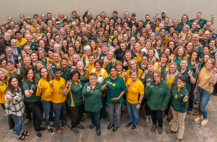A large group of NDSU Extension personnel mostly in green and gold attire pose making the "Bison Sign," one nand raised with the forefinger and pinkie finger raised to suggest bison horns. 