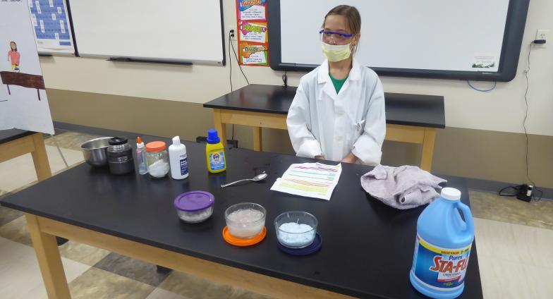 girl with mask and science lab coat on standing behind table with experiment ingredients