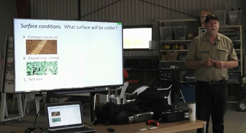 NDSU Pesticide Program specialist Andrew Thostenson, wearing a black cap, tans shirt and black pants, is standing to the right of a screen. On the screen are the words, "Surface conditions: What will be colder?"