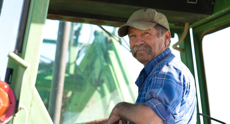 Al older farmer sits in the cab for a green tractor looking back at the camera. He has a thick, gray mustache and is wearing a tan cap and a blue plaid shirt.