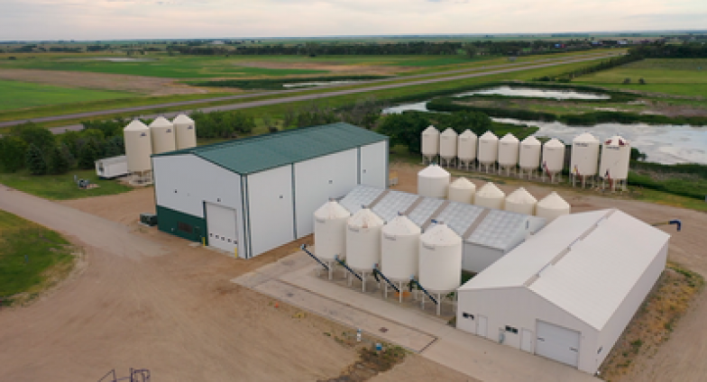 An aerial view of two large metal buildings, one all white the other white with a green roof and trim, surrounded by several, white silos.