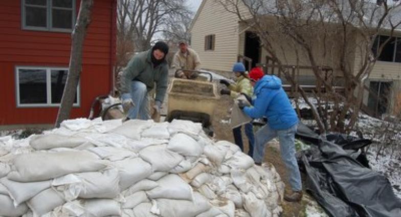 Four people in winter coats and hats work to fill white bags with sand and stack them into a dike. in front of a red house and a tan house.