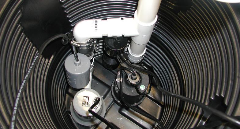 A black sump pump sits at the bottom of a hole lined with black corrugated plastic. Black electrical wires lead to the pump which is connected to white PVC pipes leading up out of the hole. 