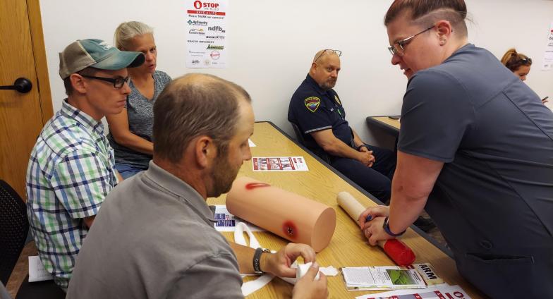 An instructor stands over a table. There are training aids that look like body parts with injuries on the table. Three people sit on the other side of the table observing.