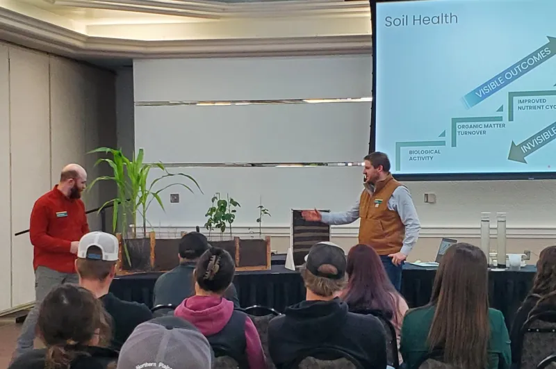 Presenters demonstrate soil health concepts with plants and soil displays to an audience in a classroom setting