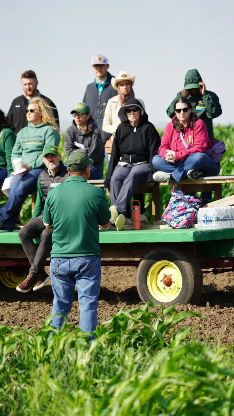 people on a trailer listening to a speaker in a crop field