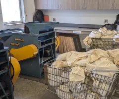 Three wire grocery carts, two full of tagged cloth bags with grain, sit near two small blue and yellow grain cleaners.