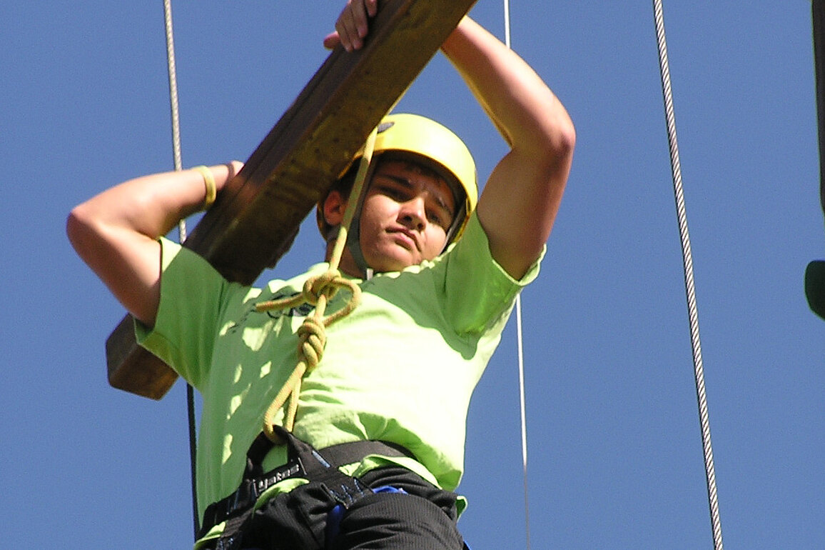 A teen wearing a bright green t-shirt, a harness, and a yellow helmet holds on to thick board as they negotiate an obstacle course.