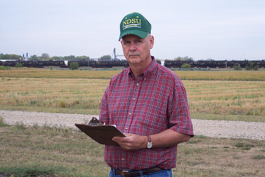 A man standing in a field wearing a baseball cap and holding a clipboard.
