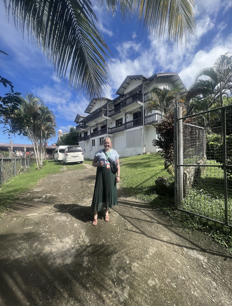 Photo of Isabel Lundberg wearing a green pleated skirt and gray shirt in front of a two-story white building with brown balconies in Fijig in Fiji