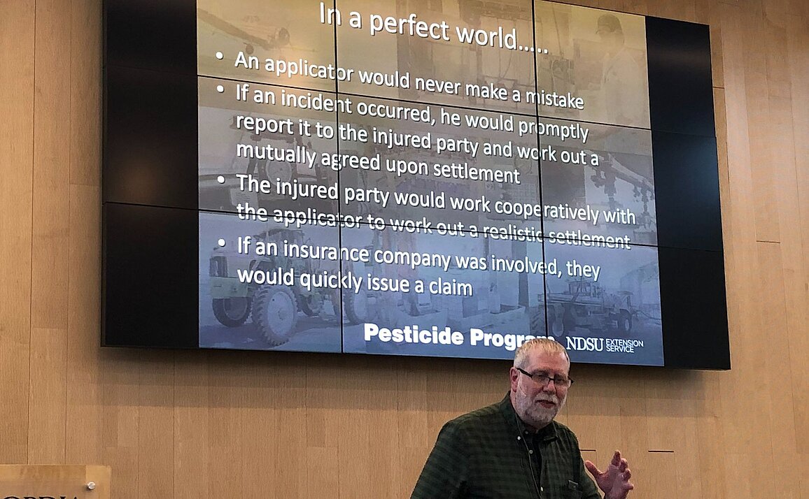 Andrew Thostenson, Extension pesticide program specialist, stands in front of a projected slide with the text, "In a perfect world....an applicator would never make a mistake; if an incident occurred, he would promptly report it to the injured party and work out a mutually agreed upon settlement; The injured party would work cooperatively with the applicator to work out a realistic settlement; if an insurance company was involved, they would quickly issue a claim.
