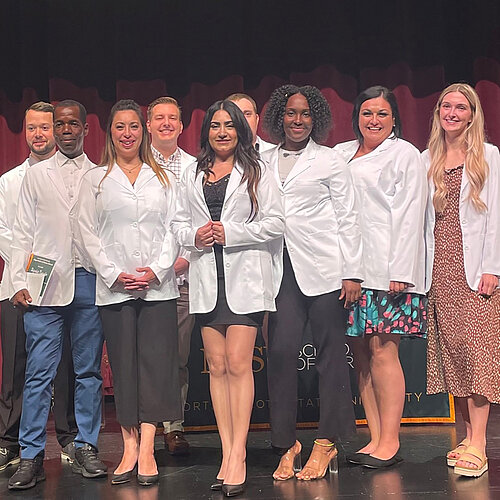 Photo of NDSU Accelerated BSN students in group wearing their white medical coats while standing on a stage at a ceremony.