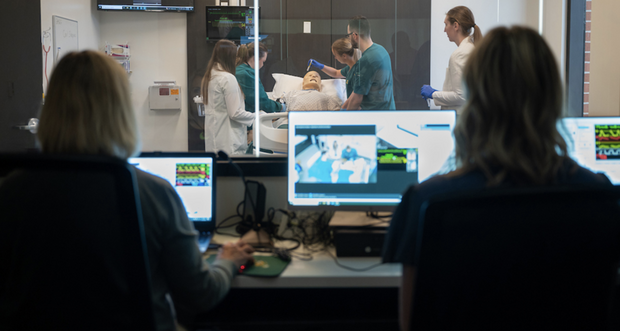 Photo of NDSU Nursing and Pharmacy students working in a Simulation Lab with a manikin that is experiencing code blue, as their professors watch and record from control room