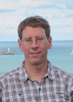 Picture of Alan Denton standing by the sea with sailboats and lighthouse in background.