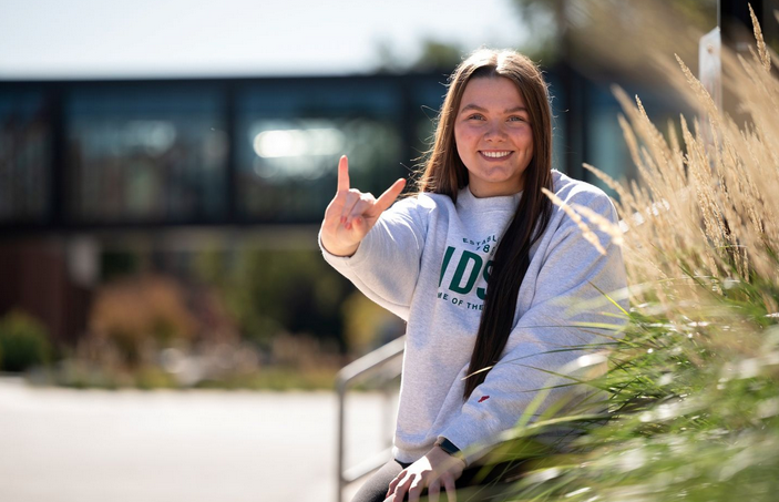 Photo of nutrition science student Masynn Janicke smiling and showing the Bison team horns up hand sign as she stands in front of a building on the NDSU campus