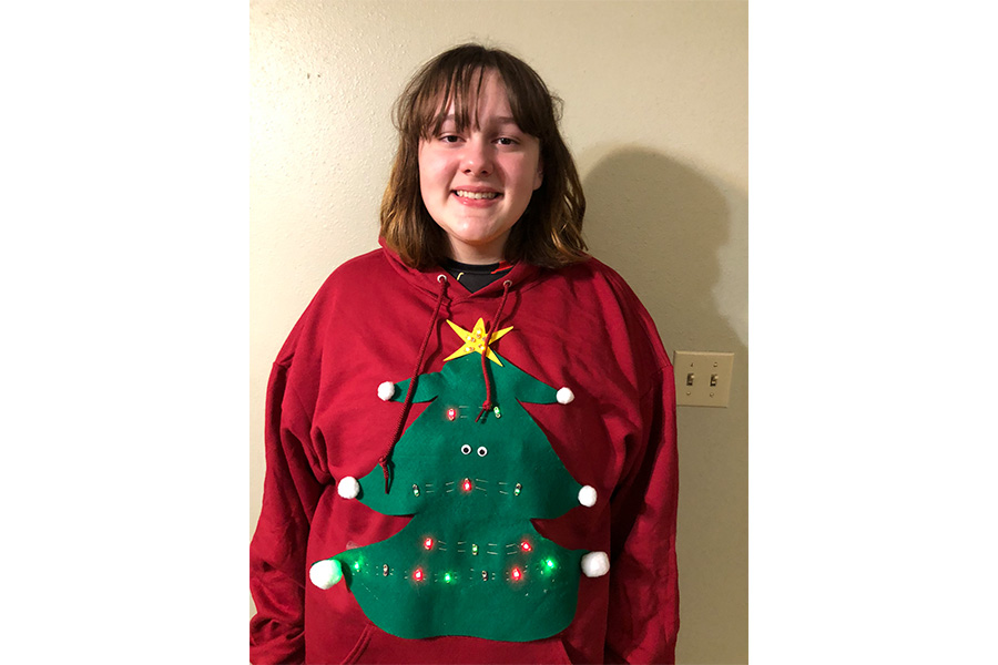 A girl stands against a wall wearing a holiday sweater. 
