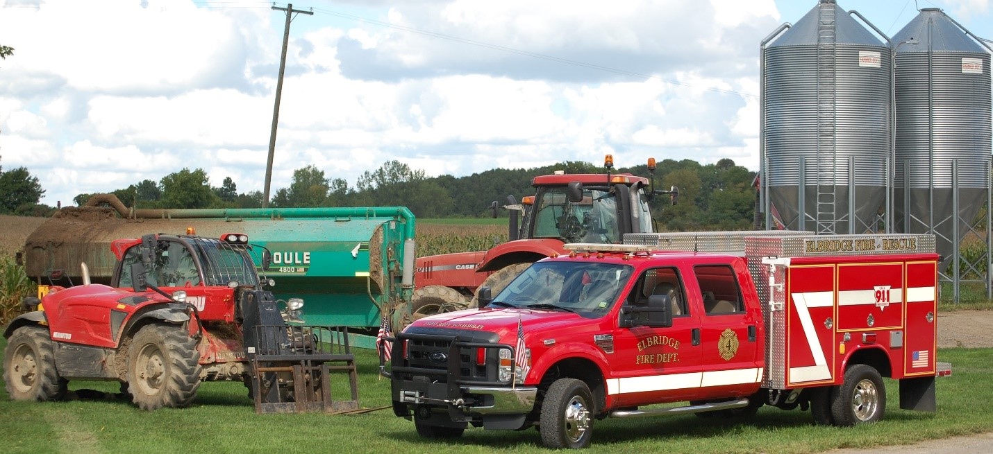 A fire engine parked next to some farm equipment with a grain silo in the background