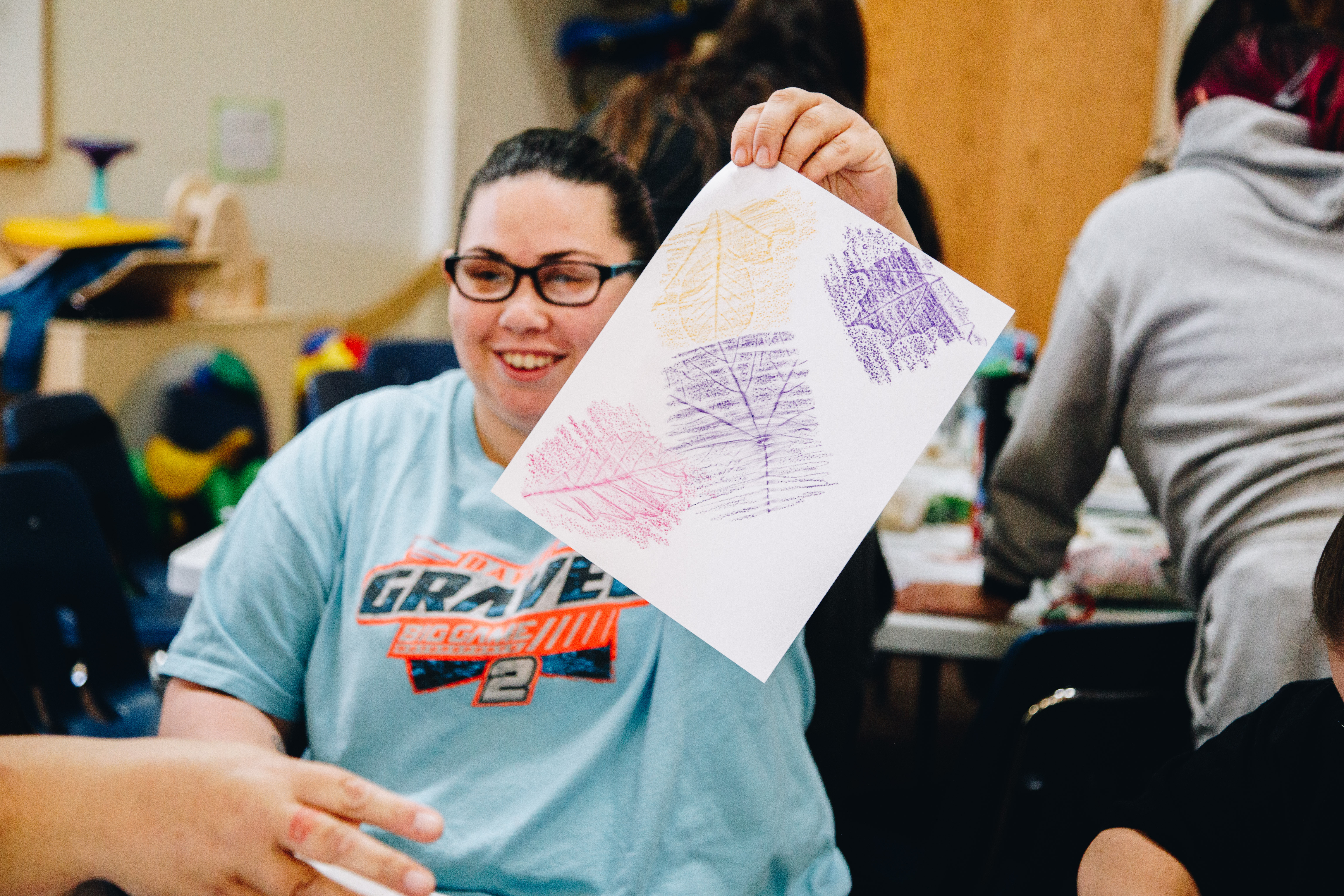 A woman in glasses holds up a piece of paper with leaf patterns made using crayons on it